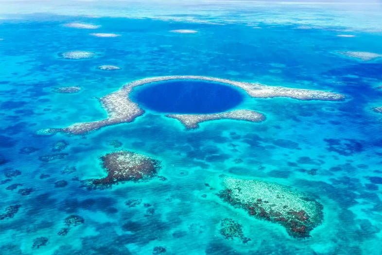 Круг темно-синей воды в океане открывает подводную пещеру, окруженную белыми рифами.